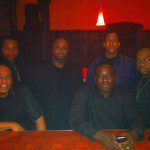 A group of men sitting at a table in front of a red wall.